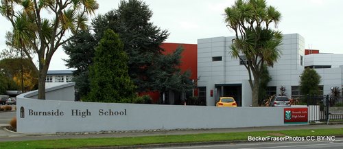 Burnside High School's front gate. Tied to the wall further down is a temporary red and green sign reading "Avonside Girls High School." It has Avonside Girls' logo on it.