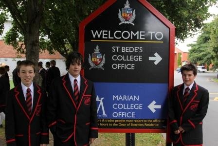 Three St Bede's boys in uniform standing beside the welcome sign at their school. The sign is split top-and-bottom into two sections. The top section is black, reads "St Bede's College Office" with their school logo and an arrow pointing to the right. The bottom half is blue, and reads "Marian College Office" with their school logo and an arrow pointing to the left.